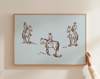 May include: A framed print featuring three brown illustrations of cowboys on horseback against a light blue background. The cowboys wear hats and are depicted in a simple, sketched style. The frame is made of light-colored wood.