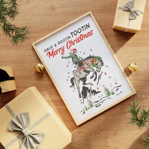 Puede incluir: Un cuadro enmarcado con el texto "Have a Rootin Tootin Merry Christmas" y un vaquero montando un caballo. El vaquero lleva un gorro de Pap&aacute; Noel. El cuadro est&aacute; rodeado de cajas de regalo y adornos navide&ntilde;os.