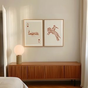 May include: Two framed prints of playing cards, one with an ace of spades and the other with a cowboy riding a bucking horse. The prints are hanging on a white wall above a wooden cabinet with a lamp on top.