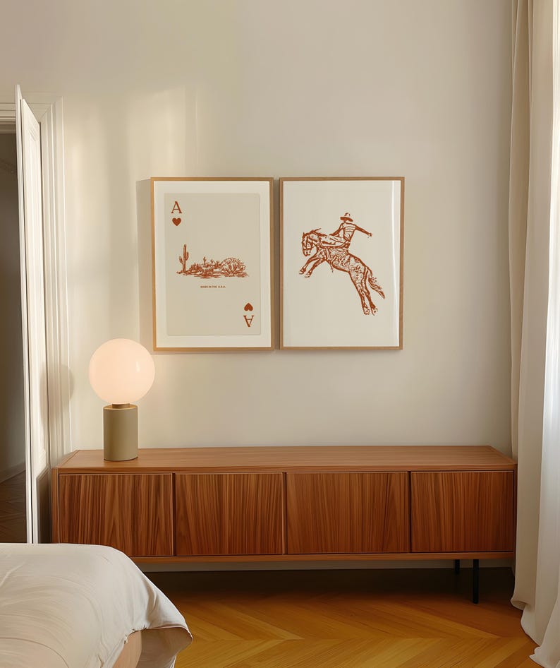 May include: Two framed prints of playing cards, one with an ace of spades and the other with a cowboy riding a bucking horse. The prints are hanging on a white wall above a wooden cabinet with a lamp on top.