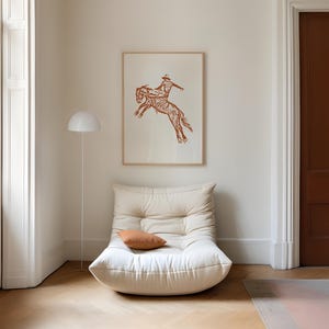 May include: A white, tufted armchair with a brown pillow sits in a room with a framed print of a brown silhouette of a cowboy riding a horse. The room has white walls and a wooden floor.