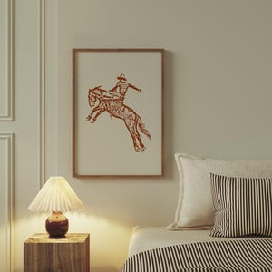 May include: A framed print of a brown line drawing of a cowboy riding a bucking horse. The print is hanging on a wall above a bed with a striped comforter.