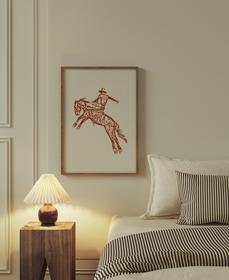 May include: A framed print of a brown line drawing of a cowboy riding a bucking horse. The print is hanging on a wall above a bed with a striped comforter.