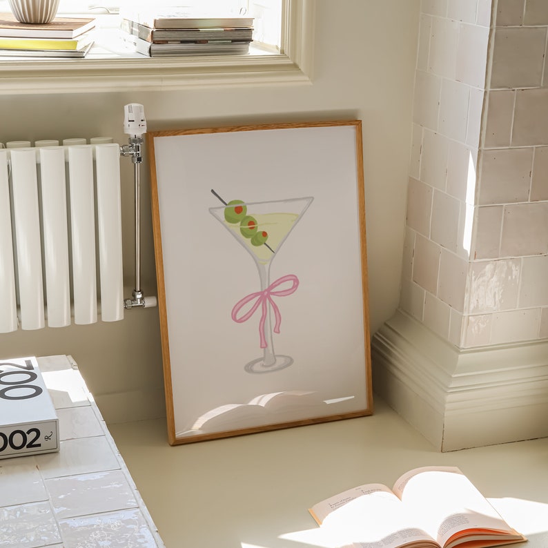 May include: A framed art print showing a martini cocktail illustration. The artwork features a martini glass with a yellow liquid, green olives on a cocktail stick, and a pink bow. The frame is made of light brown wood.