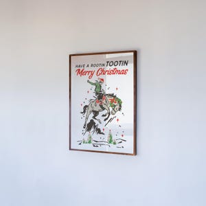 Puede incluir: Un estampado enmarcado con tem&aacute;tica navide&ntilde;a. La obra de arte presenta a un vaquero montando un caballo que se encabrita, con el texto "Have a Rootin' Tootin' Merry Christmas" en rojo y verde. El estampado est&aacute; en un marco de madera marr&oacute;n.