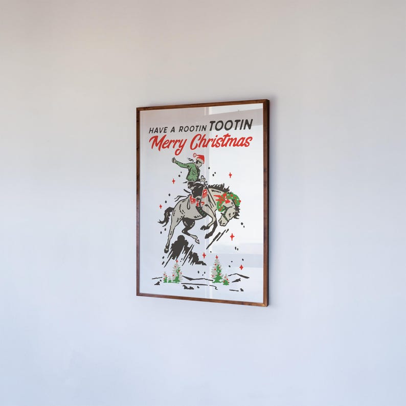 Puede incluir: Un estampado enmarcado con tem&aacute;tica navide&ntilde;a. La obra de arte presenta a un vaquero montando un caballo que se encabrita, con el texto "Have a Rootin' Tootin' Merry Christmas" en rojo y verde. El estampado est&aacute; en un marco de madera marr&oacute;n.