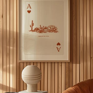 May include: A framed print of an ace of spades playing card with a desert landscape illustration. The card is printed in red on a white background with the text "Made in the U.S.A.". The print is hanging on a wood panel wall.