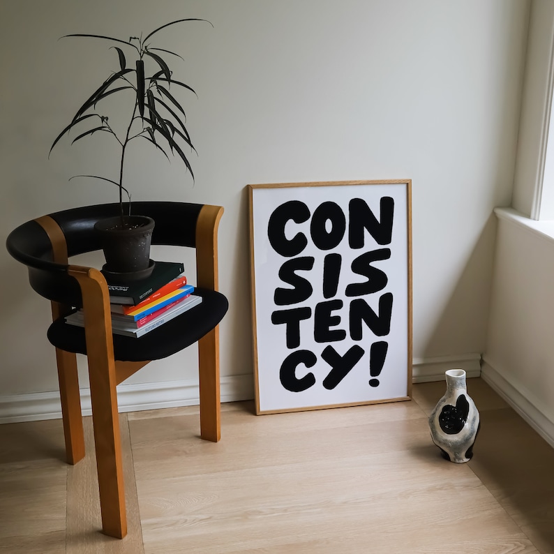 May include: A black and white poster with the word "CONSISTENCY" in large, bold, black letters. The poster is framed in a light wood frame and is hanging on a white wall. A small potted plant and a wooden chair are in the foreground.