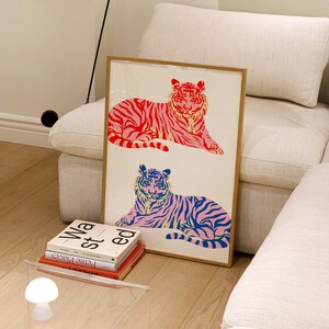 May include: A framed print featuring two stylised tigers, one in pink and red and the other in blue and pink. The tigers are lying down and facing the viewer. The print is on a white background and has a wooden frame.