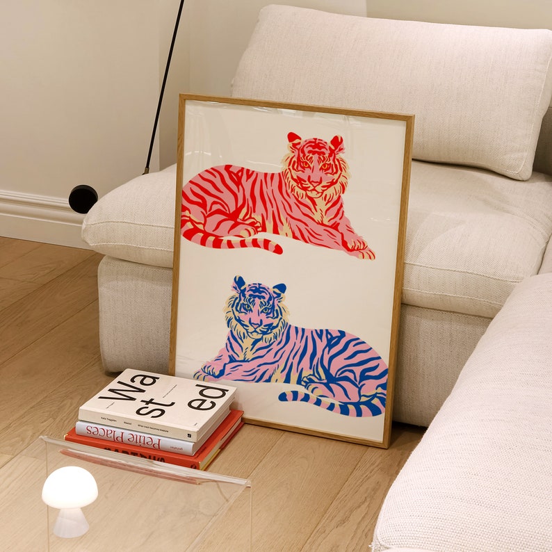 May include: A framed print featuring two stylised tigers, one in pink and red and the other in blue and pink. The tigers are lying down and facing the viewer. The print is on a white background and has a wooden frame.