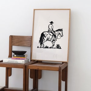 Op de afbeelding: Een zwart-witte lijntekening van een cowboy die op een paard rijdt. De tekening is ingelijst in een licht houten lijst en hangt aan een witte muur.