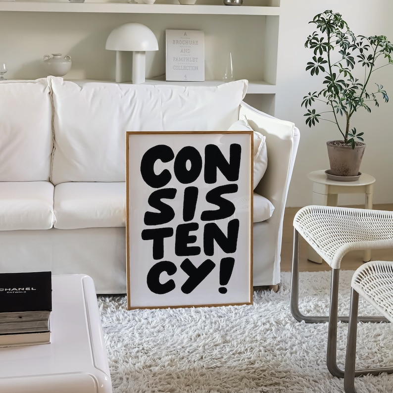 May include: A framed poster with the word "CONSISTENCY" in large, black, bold letters. The letters are stylized and slightly overlapping. The poster is hanging on a white wall in a living room setting.