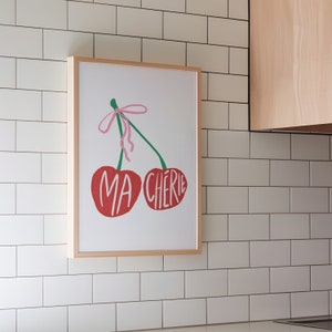 May include: A framed print of two red cherries with green stems and a pink bow. The cherries are labeled "MA CHERIE" in white text.