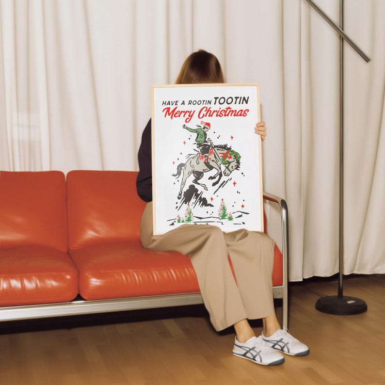 Puede incluir: Un p&oacute;ster enmarcado con el texto "Have a Rootin Tootin Merry Christmas" sobre un gr&aacute;fico de un vaquero montando un caballo. El vaquero lleva un gorro de Pap&aacute; Noel. El p&oacute;ster lo sostiene una persona sentada en un sof&aacute; rojo.