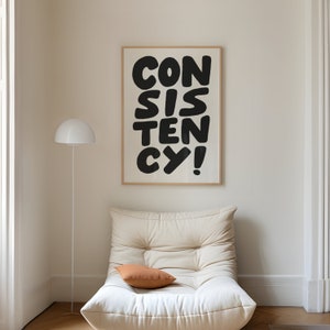 May include: A white framed poster with black text that reads "CONSISTENCY!" hanging on a white wall above a white couch with a brown pillow.