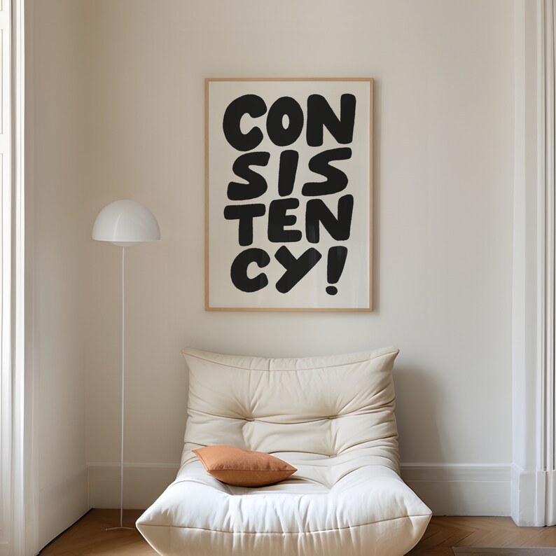 May include: A white framed poster with black text that reads "CONSISTENCY!" hanging on a white wall above a white couch with a brown pillow.