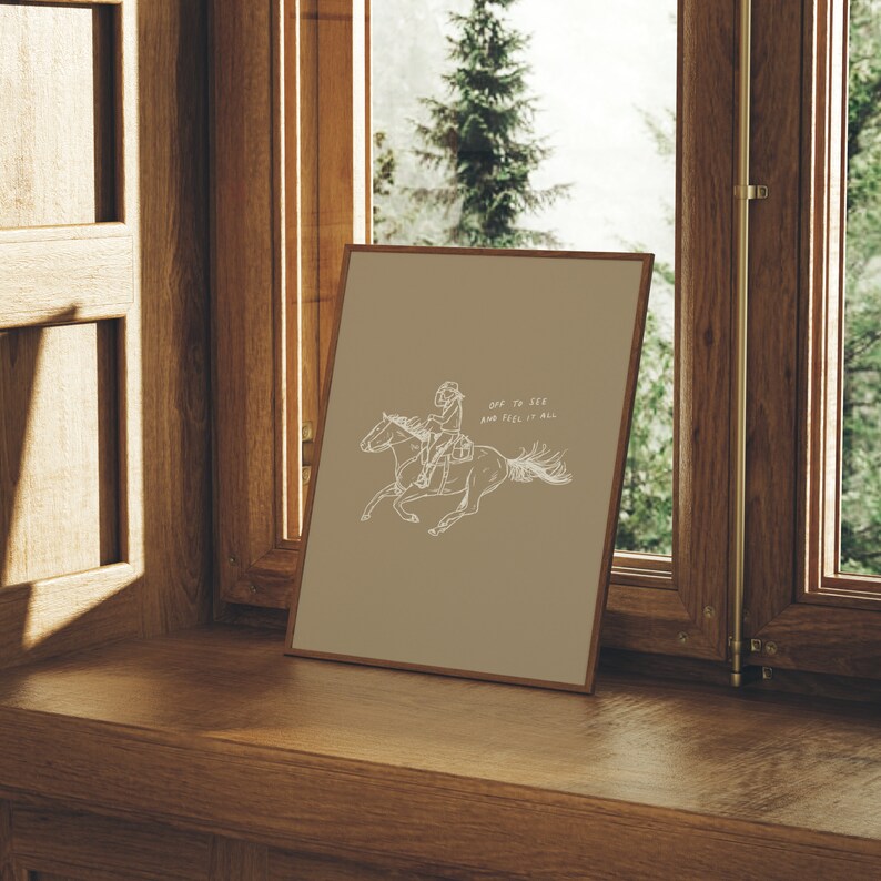 Pu&ograve; includere: Una stampa incorniciata con uno sfondo marrone e un disegno a linea bianca di una persona a cavallo. Il testo "Off to see and feel it all" &egrave; scritto in bianco sulla stampa.