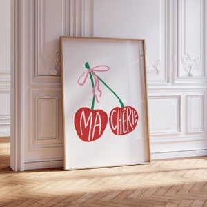 May include: A framed print with a white background featuring two red cherries with green stems and a pink ribbon. The cherries have the words "MA CHERIE" written on them.