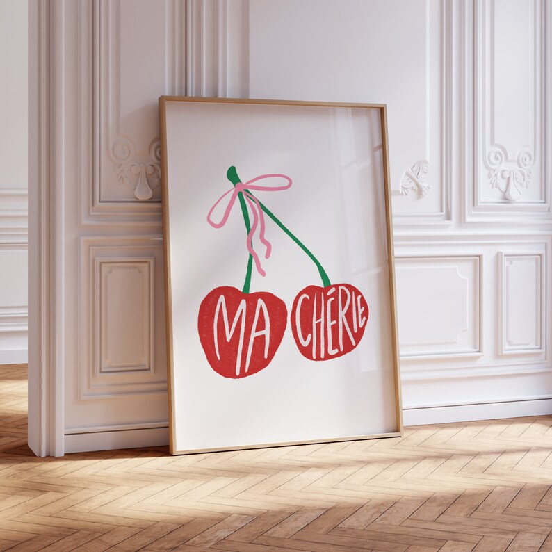 May include: A framed print with a white background featuring two red cherries with green stems and a pink ribbon. The cherries have the words "MA CHERIE" written on them.
