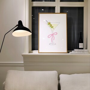 May include: A framed print depicting a martini glass with olives and a pink bow. The artwork is displayed on a white windowsill, next to a black lamp and a dark bottle. The background includes a window with closed blinds.