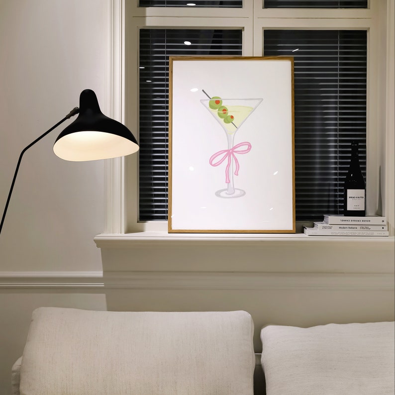 May include: A framed print depicting a martini glass with olives and a pink bow. The artwork is displayed on a white windowsill, next to a black lamp and a dark bottle. The background includes a window with closed blinds.