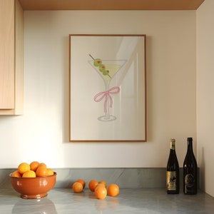 May include: A framed print depicting a martini glass with olives and a pink bow. The artwork is above a countertop with a bowl of oranges and two wine bottles. The color scheme is neutral with accents of green and pink.