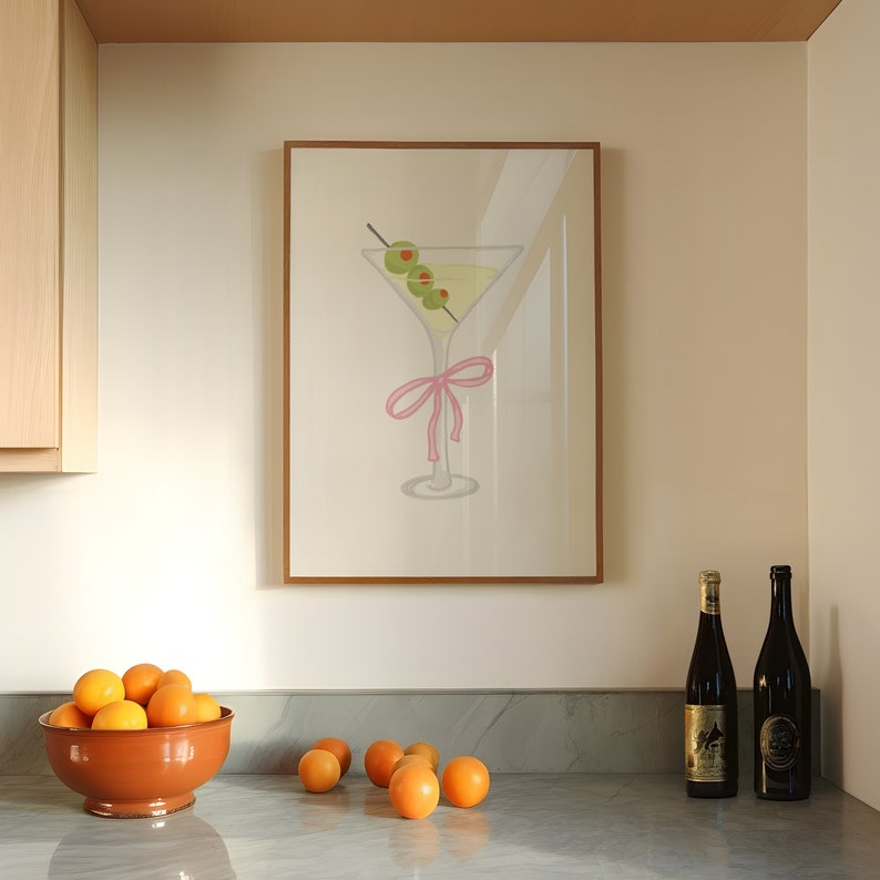 May include: A framed print depicting a martini glass with olives and a pink bow. The artwork is above a countertop with a bowl of oranges and two wine bottles. The color scheme is neutral with accents of green and pink.