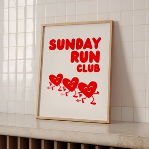 May include: A white framed print with the text "SUNDAY RUN CLUB" in red letters.  Four red hearts with smiling faces are running across the bottom of the print.