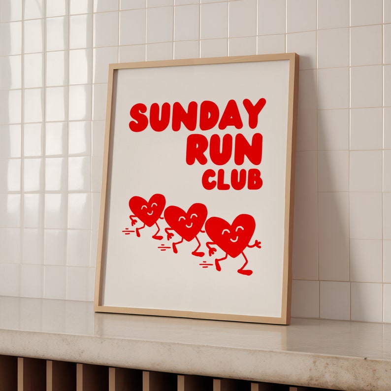 May include: A white framed print with the text "SUNDAY RUN CLUB" in red letters.  Four red hearts with smiling faces are running across the bottom of the print.
