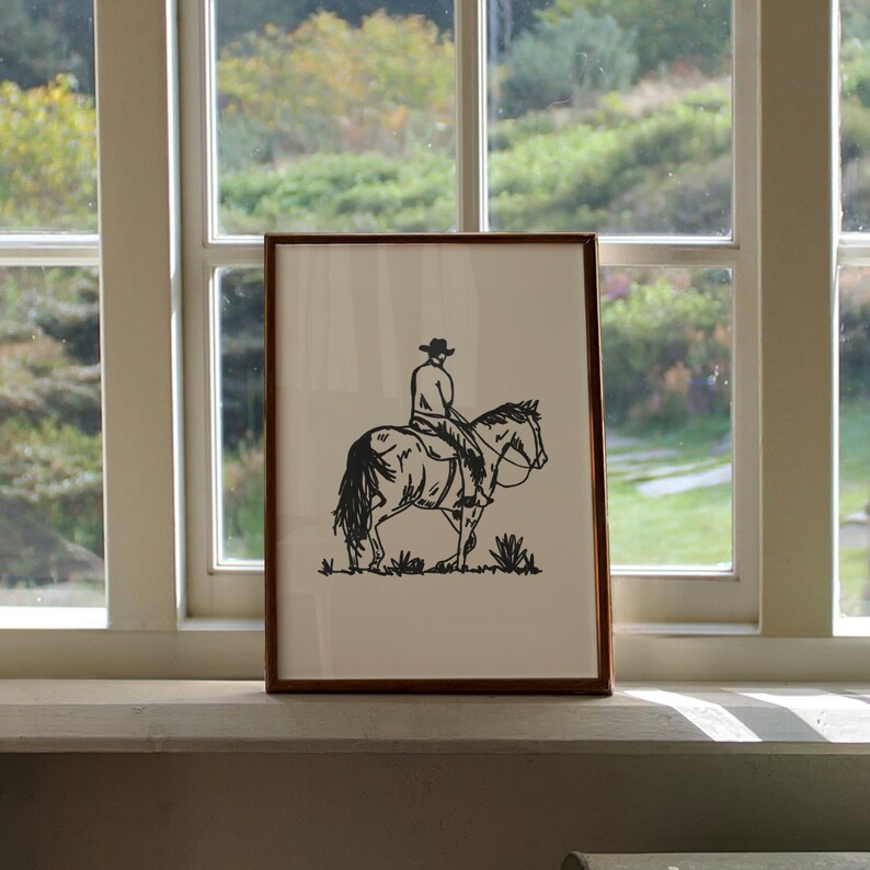 Op de afbeelding: Een zwart-witte lijntekening van een cowboy die op een paard rijdt. De tekening is ingelijst in een bruin lijst en hangt aan een muur.