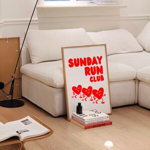May include: A white couch with a framed poster that says "SUNDAY RUN CLUB" in red letters. The poster features five red hearts with faces running. There are books stacked below the poster.