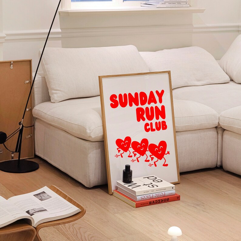 May include: A white couch with a framed poster that says "SUNDAY RUN CLUB" in red letters. The poster features five red hearts with faces running. There are books stacked below the poster.