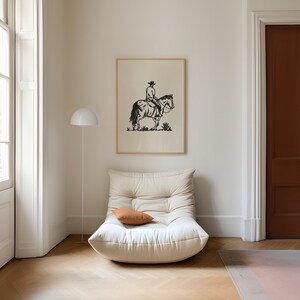 Op de afbeelding: Een witte fauteuil met een bruin kussen voor een ingelijst zwart-witte afbeelding van een cowboy die een paard berijdt. De fauteuil staat in een kamer met witte muren en een houten vloer.