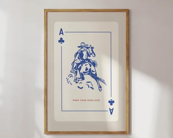 May include: Framed art print featuring a blue illustration of a cowboy riding a bucking bronco on an Ace of Clubs playing card. The text "MAKE YOUR OWN LUCK" is printed below the illustration. The print is in a wooden frame.