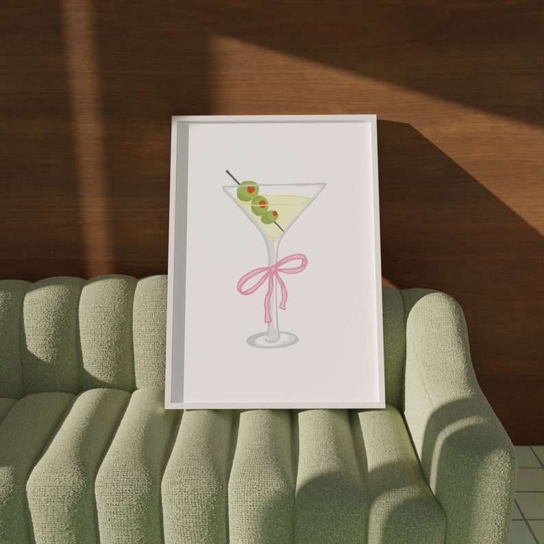 May include: A framed print depicting a cartoon martini glass with olives on a cocktail stick. The glass is decorated with a pink bow. The artwork is set against a light grey background.