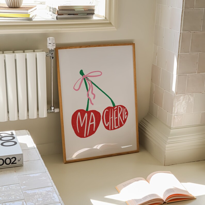 May include: A framed print with a white background and two red cherries with green stems and pink ribbons. The cherries have the words "Ma Cherie" written on them.