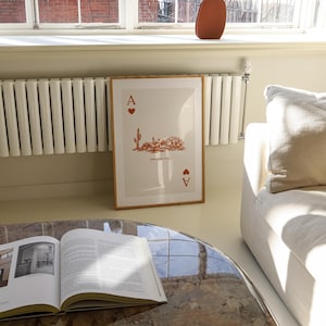 May include: A framed print of an Ace of Spades playing card with a desert landscape design in orange. The card is framed in a light wood frame and is hanging on a wall in a room with a white radiator and a white couch.