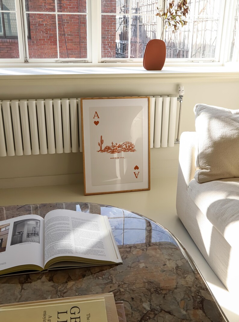 May include: A framed print of an Ace of Spades playing card with a desert landscape design in orange. The card is framed in a light wood frame and is hanging on a wall in a room with a white radiator and a white couch.