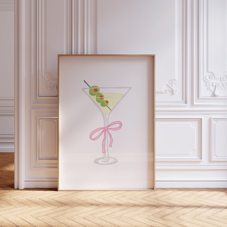May include: Framed print with a watercolour illustration of a martini. The cocktail glass contains a yellow liquid and is garnished with three green olives on a cocktail stick. A pink bow decorates the glass stem.