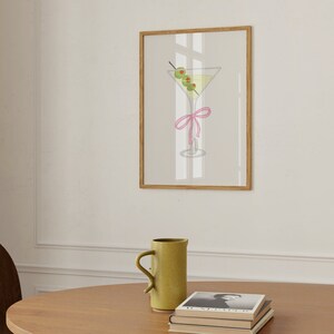 May include: A framed print showing a martini glass with olives and a pink ribbon. The cocktail has a light yellow liquid and three green olives on a cocktail stick. The frame is a light brown colour.