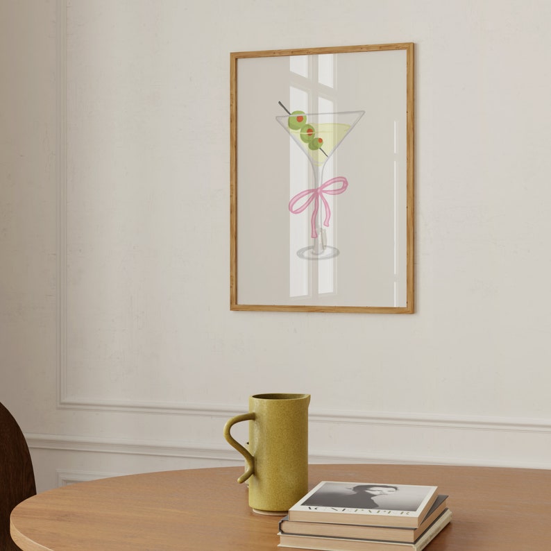 May include: A framed print showing a martini glass with olives and a pink ribbon. The cocktail has a light yellow liquid and three green olives on a cocktail stick. The frame is a light brown colour.