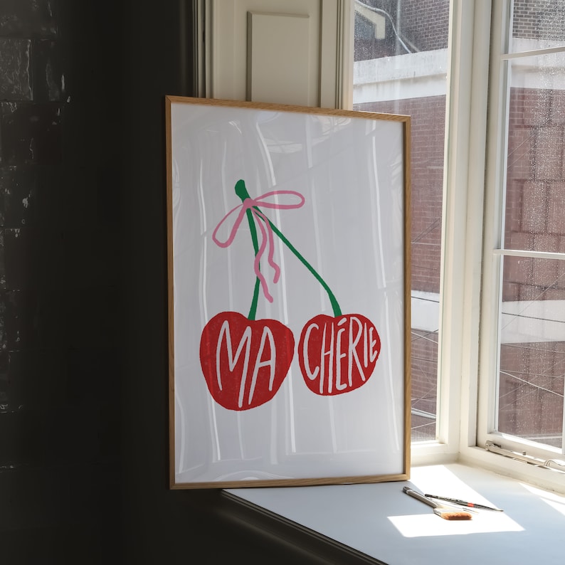 May include: A framed print with a white background featuring two red cherries with green stems and a pink ribbon. The cherries have the words "MA CHERIE" written on them.