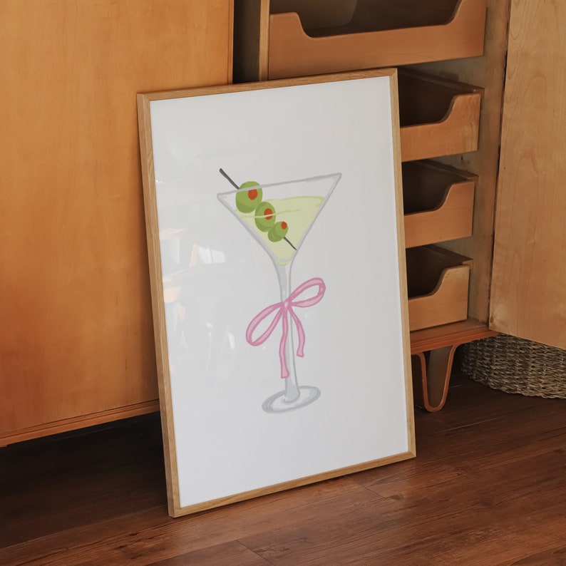 May include: A framed art print depicting a martini glass with olives on a cocktail stick and a pink bow. The glass contains a yellow liquid. The artwork is set against a white background.