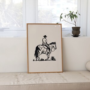 Op de afbeelding: Zwart-witte lijntekening van een cowboy die op een paard rijdt. De afbeelding is ingelijst en hangt aan een muur.