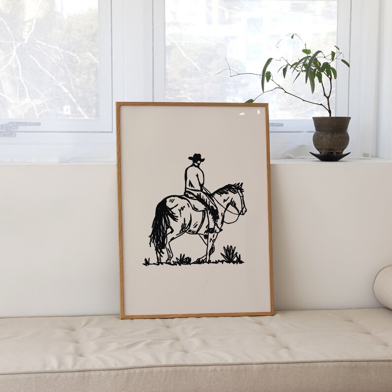 Op de afbeelding: Zwart-witte lijntekening van een cowboy die op een paard rijdt. De afbeelding is ingelijst en hangt aan een muur.