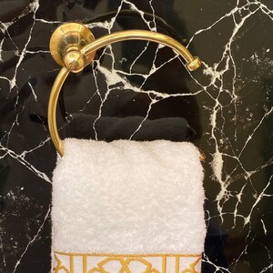 May include: White towel with a gold embroidered geometric pattern hanging on a gold towel ring against a black marble background.