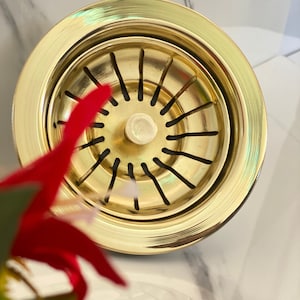 May include: A gold-toned metal sink strainer with a circular design and a central drain hole. The strainer has a series of evenly spaced slots around the edge.
