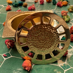 Puede incluir: Un quemador de incienso de metal dorado con un acabado martillado decorativo y una tapa circular calada. La tapa tiene una serie de cuadrados recortados alrededor del borde.