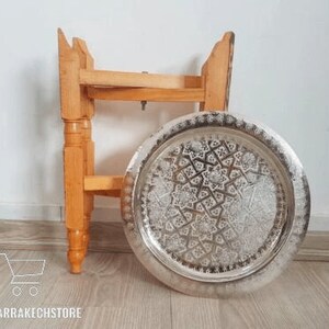 May include: A wooden stand with a round, silver tray. The tray has an intricate, embossed design.