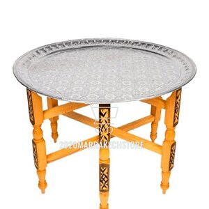 May include: A round silver tray table with a decorative pattern on the tray. The table has a wooden base with intricate carvings and four legs.